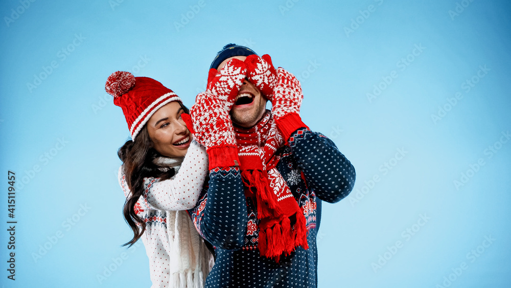 Cheerful woman in sweater and mittens covering eyes of boyfriend on blue background