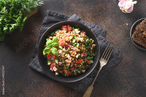 Fotografie Cooked whole grain cereal spelt as salad with vegetables, tomatoes, cucumber, greens on brown background
