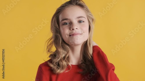 Beautiful cute blond teenager girl with wavy hair dressed in red sweater posing on camera over yellow background