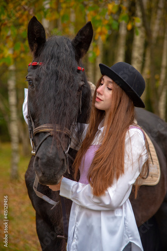 A girl stands next to a horse on the background of an autumn landscape.