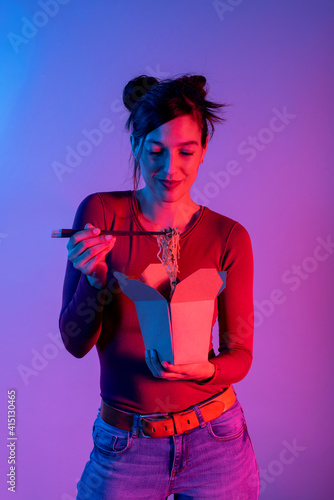 Attractive woman eating Asian noodles for lunch with chopsticks in studio with blue and red lights