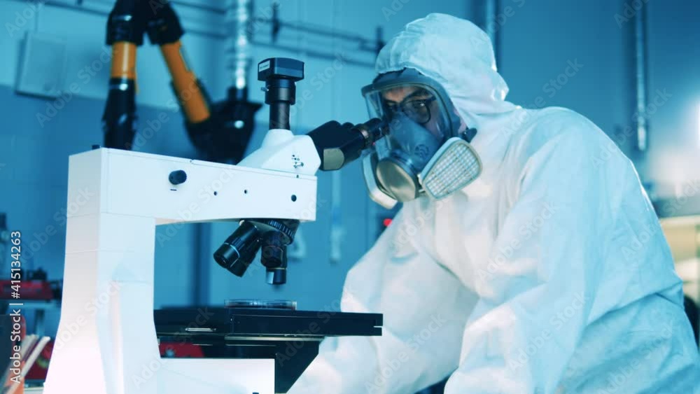 Male specialist in a hazmat suit is operating a microscope
