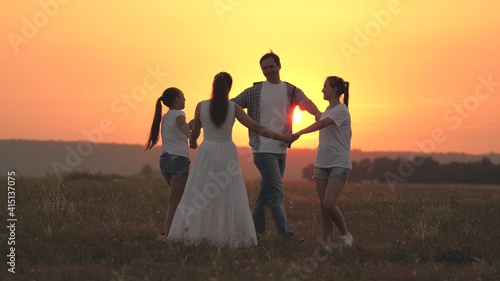 A happy family, a young mother, dad and daughters play and dance together in the sun, have fun in field. Children with parents play in park on grass at sunset. Happy family childhood and health