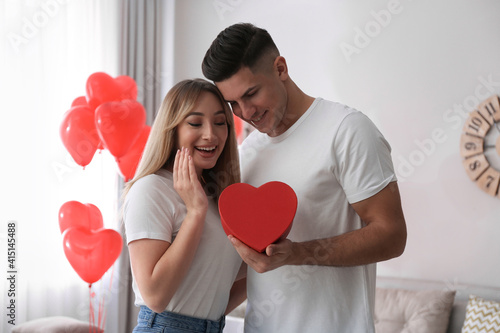 Man presenting gift to his girlfriend in room decorated with heart shaped balloons. Valentine's day celebration