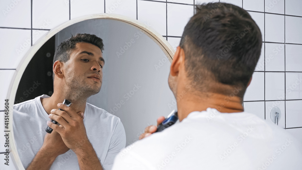 Obraz premium young man shaving neck with electric razor near mirror in bathroom, blurred foreground