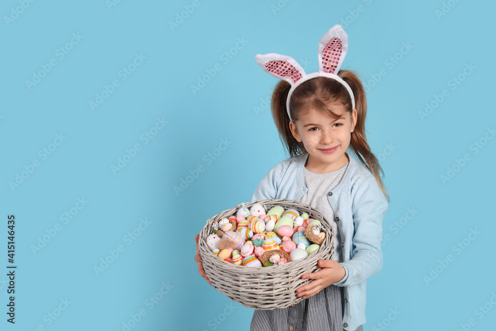 Happy little girl with bunny ears holding wicker basket full of Easter eggs on light blue background. Space for text Happy little girl with bunny ears holding wicker basket full of Easter eggs on light blue background. Space for text