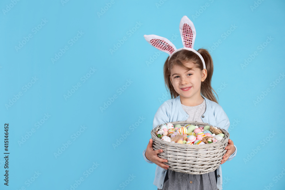 Happy little girl with bunny ears holding wicker basket full of Easter eggs on light blue background. Space for text Happy little girl with bunny ears holding wicker basket full of Easter eggs on light blue background. Space for text