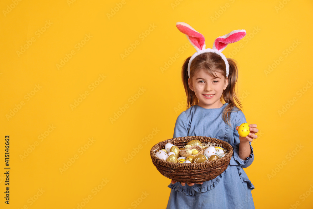 Happy little girl with bunny ears holding wicker basket full of Easter eggs on orange background. Space for text