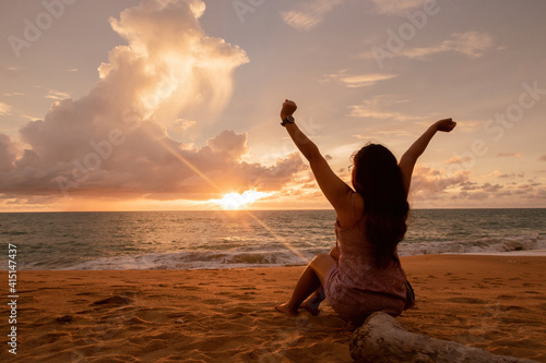 Concept of tourism and relaxation, an woman sitting on a piece of wood on the beach looking at the sky and the sea at sunset, expressing freedom and relaxation, raising her hands, admiring nature.
