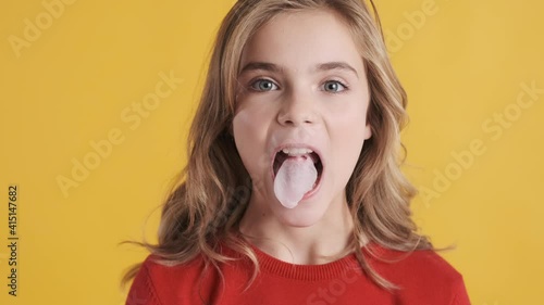 Funny blond teenager girl blowing big bubble from chewing gum on camera isolated on yellow background. Candy bubble photo