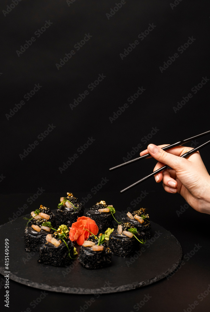 Hand with chopsticks wants to take custom sushi roll with black rice, crab meat, avocado, smoked salmon mousse, oar caviar, masago, shrimp cocktail, edible gold leaf, ginger, wasabi on black table. Hand with chopsticks wants to take custom sushi roll with black rice, crab meat, avocado, smoked salmon mousse, oar caviar, masago, shrimp cocktail, edible gold leaf, ginger, wasabi on black table.