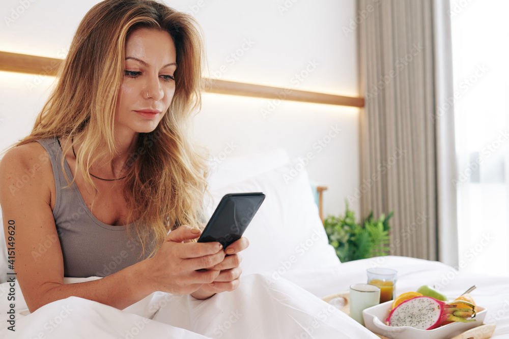 Attractive serious young woman sitting in bed, eating breakfast and checking social media and answering text messages on smartphone