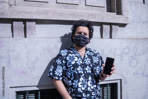 an adult influencer with long hair wearing a protraction mask at street using a mobile phone and sharing with followers as content creator at city in virus time, close up portrait photo