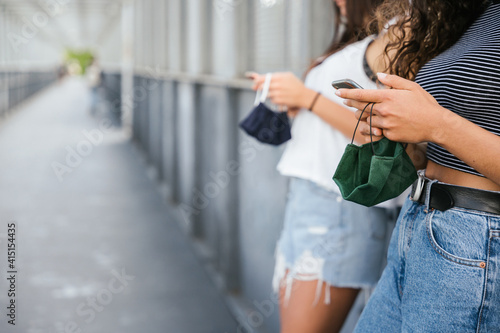 Portrait two young girls using their smartphones leaning against wall of iron bridge with face masks in hands during Coronavirus Covid-19 pandemic Millennials chat on social networks and watch media