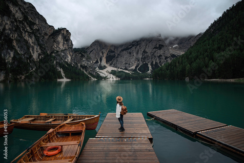 Young traveler girl with hat and vintage backpack on jetty of lago di Braies surrounded by the mountains of the Italian alps 