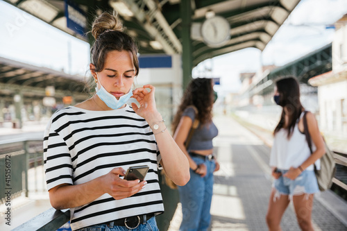 Three beautiful young female friends at the station wait for the train for their vacation during Coronavirus Covid-19 pandemic wearing face masks - Millennials use smartphone and others talk together