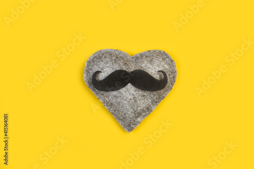 Corazon gris de fieltro con un bigote mostacho negro sobre un fondo amarillo brillante liso y aislado. Vista superior. Copy space