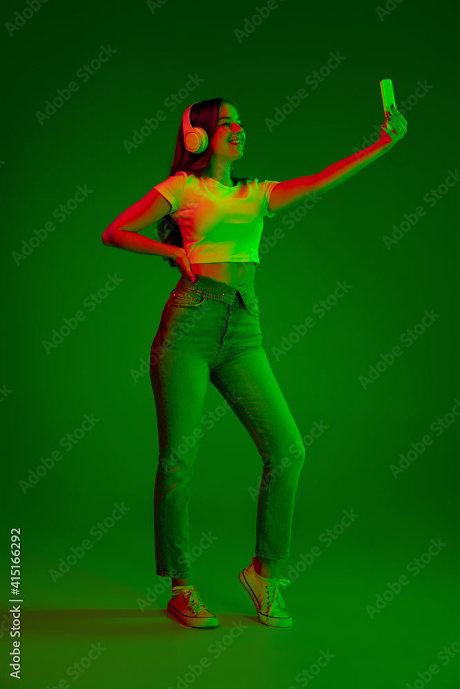 Caucasian woman's portrait isolated on green studio background in multicolored neon light. Taking selfie in headphones. Concept of human emotions, facial expression, sales, ad, fashion. Copyspace. Caucasian woman's portrait isolated on green studio background in multicolored neon light. Taking selfie in headphones. Concept of human emotions, facial expression, sales, ad, fashion. Copyspace.