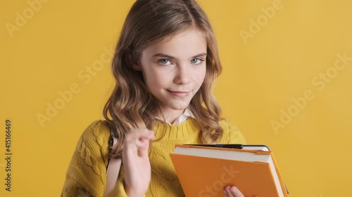 Beautiful blond teenage student girl dressed in sweater standing with books playfully looking in camera over yellow background