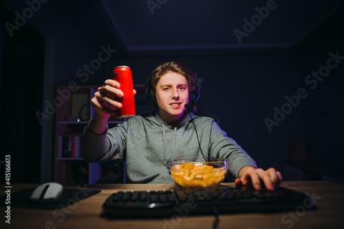 Positive young male gamer sitting in a room with purple light eating chips and holding a jar of cola  looking at the camera and smiling. Guy in the headset streams and eats snacks