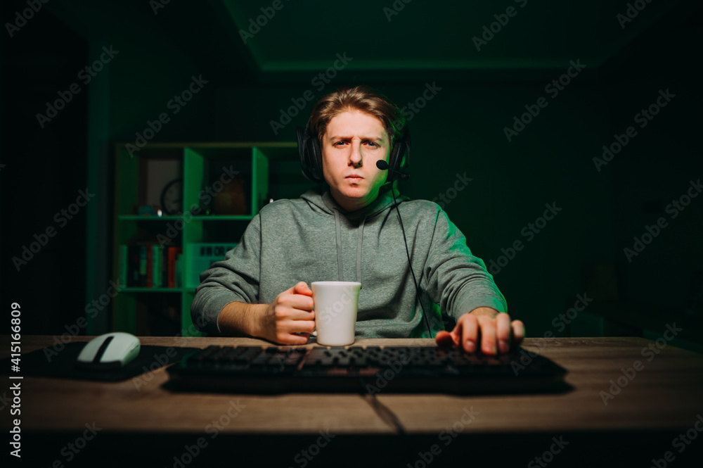 Portrait of a pensive gamer in a headset on his head uses a computer with a serious face and drinks tea. Guy at night in the bedroom plays computer games. Portrait of a pensive gamer in a headset on his head uses a computer with a serious face and drinks tea. Guy at night in the bedroom plays computer games.