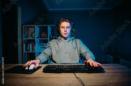 Tired young e-sportsman in a suit sits at home in a room with a blue light and trains in video games, looking at the camera with a serious face. E-sports training.