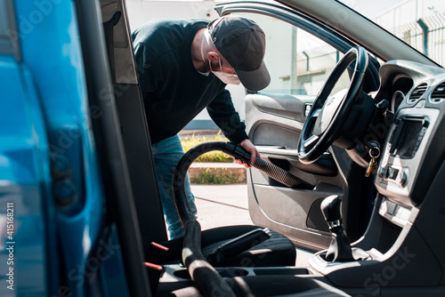 Hombre con gorra y mascarilla limpiando el interior de la puerta de un automóvil con una aspiradora profesional. Trabajador de lavado de autos