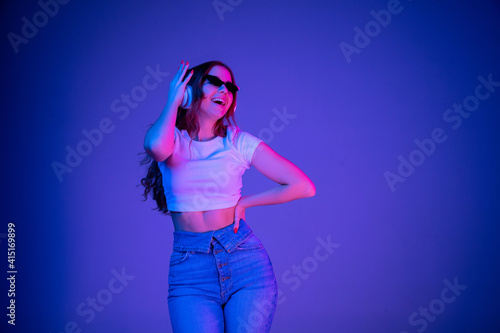 Caucasian woman's portrait isolated on blue studio background in multicolored neon light. Beautiful model with headphones. Concept of human emotions, facial expression, sales, ad, fashion. Copyspace.