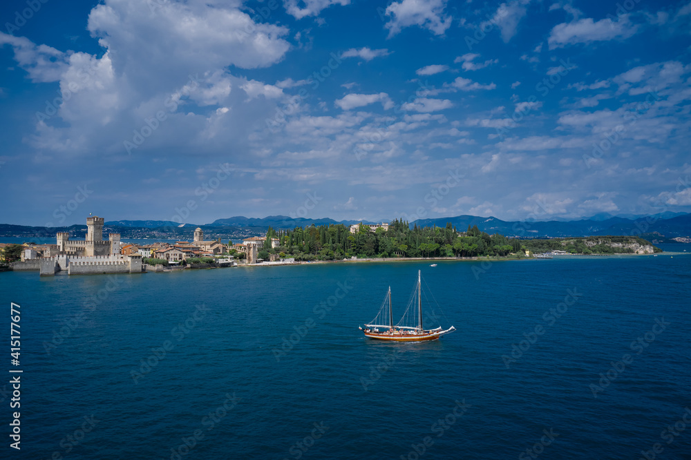The Old Sailing Ship is sailing near Sirmione Castle. Rocca Scaligera Castle in Sirmione Lake Garda Italy. Aerial view. Clouds in the blue sky.