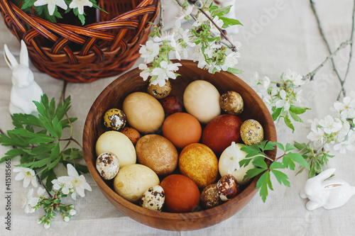 Happy Easter! Easter eggs in wooden bowl on rustic table with bunny, basket and spring flowers.