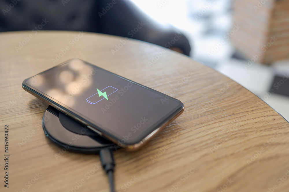 Charging mobile phone battery with wireless charging device in the table. Smartphone charging on a charging pad. Mobile phone near wireless charger Modern lifestyle concept. Charging mobile phone battery with wireless charging device in the table. Smartphone charging on a charging pad. Mobile phone near wireless charger Modern lifestyle concept.