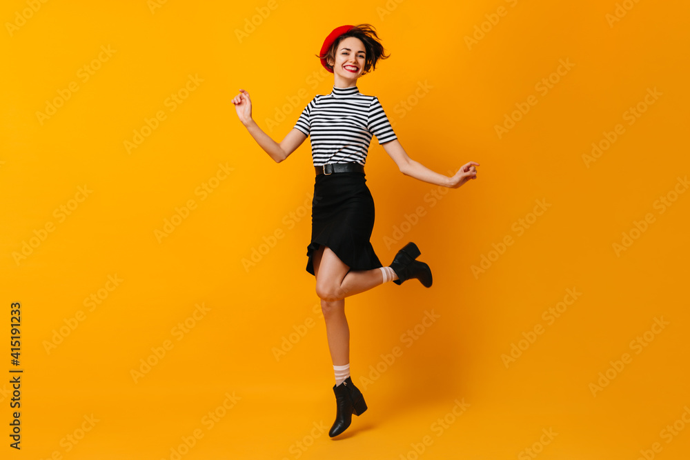 Cheerful stylish french woman dancing on yellow background. Cute young lady in beret and skirt jumping with smile.