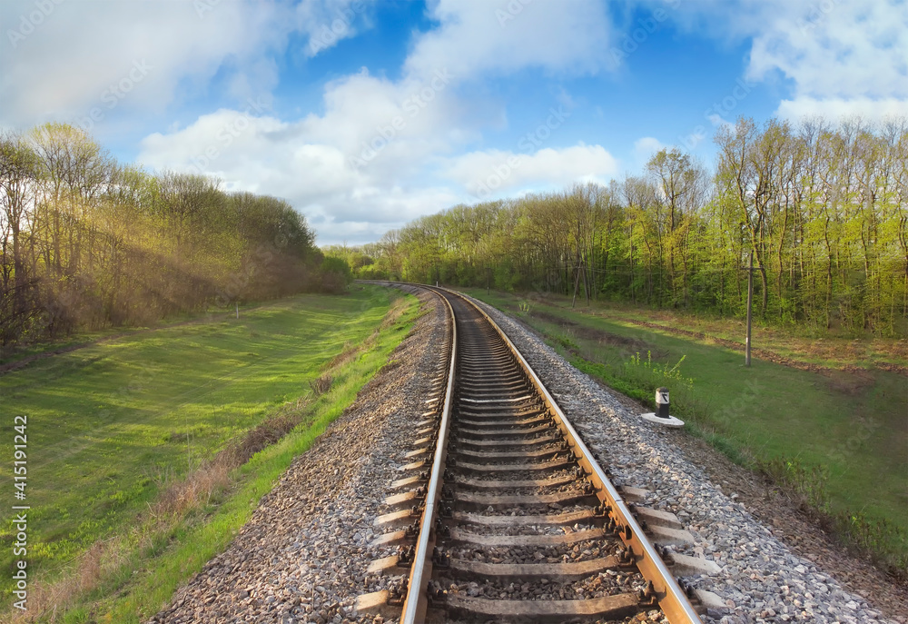 Railroad tracks with the morning sun shining through the branches of trees. Early spring, nature along the road and clouds in the blue sky. Railroad tracks with the morning sun shining through the branches of trees. Early spring, nature along the road and clouds in the blue sky.