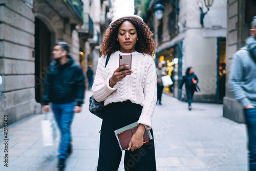 Serious dark skinned woman in trendy wear walking on street share content browsing web pages on smartphone,beautiful 20s african american female checking notification on mobile phone of banking app