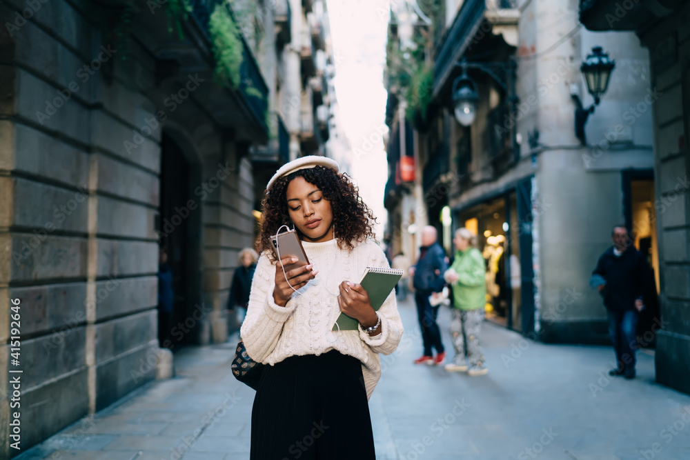Fototapeta premium Pensive attractive dark skinned woman in trendy wear standing on city street reading messages and news on smartphone,beautiful african american millennial female dialing number on cellular for calling