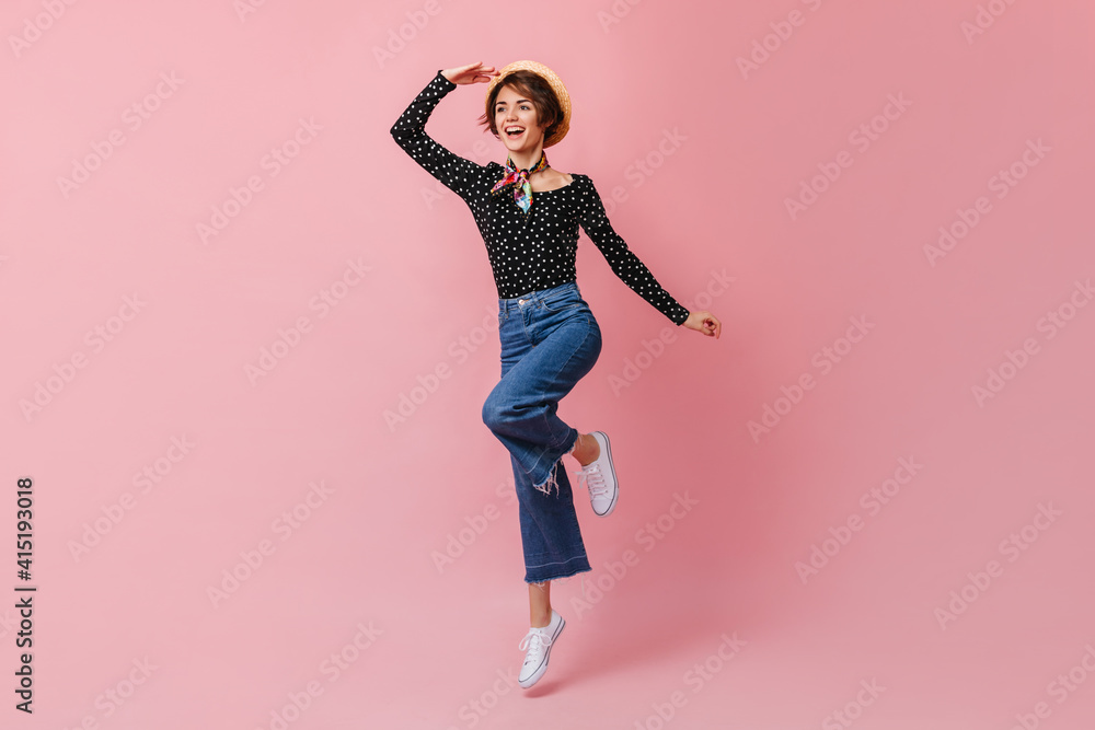 Laughing woman in straw hat looking in distance. Studio shot of amazing girl in vintage pants dancing on pink background.