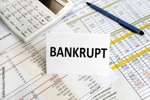 The word bankrupt is written on a white sheet that stands on a financial document near a white calculator and a blue pen. Business and financial concept