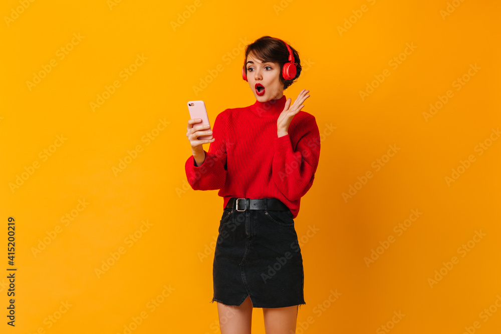 Shocked young lady in black skirt looking at smartphone. Studio shot of emotional woman in headphones.