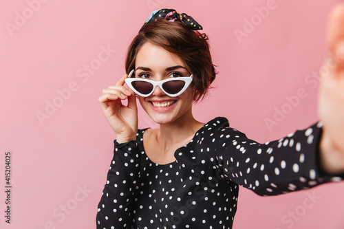 Glad girl in sunglasses taking selfie with smile. Studio shot of blissful woman with short haircut posing on pink background.