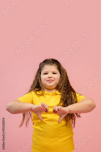 Thumbs down. Happy, smiley little caucasian girl isolated on coral pink studio background with copyspace for ad. Looks happy, cheerful. Childhood, education, human emotions, facial expression concept.