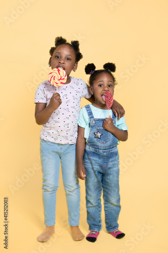 Full length shot of two cute little African girls licking sweet colorful lollipops, smiling to camera on yellow color background