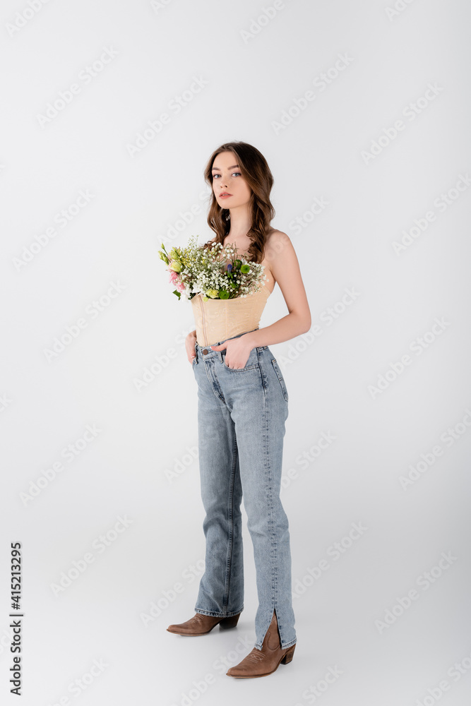 Young model with flowers in blouse and hands in pockets of jeans looking at camera on grey background Young model with flowers in blouse and hands in pockets of jeans looking at camera on grey background