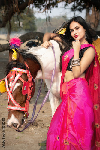 Outdoor portrait of very beautiful Indian girl wearing saree and holds the reins saddled horse and posing fashionable in blurred background. Lifestyle and Fashion.