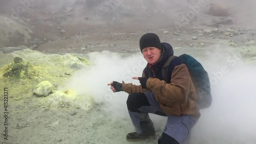 Caucasian man sits by active fumarole with sulfur surface in crater of Mutnovsky volcano on Kamchatka Peninsula, Russia. At some point, the smoke from the fumarole moves towards the person. photo