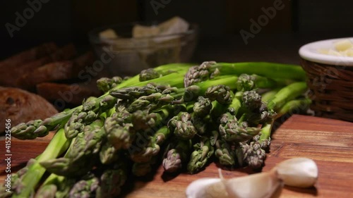 Peeled asparagus spear falls on the bunch of asparagus on a wooden board next to tha garlic and cheese photo