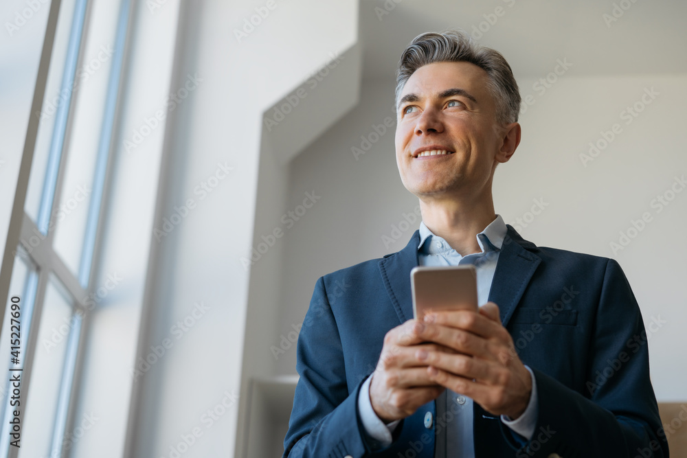 Handsome mature businessman holding mobile phone looking at window standing in modern office. Successful business concept Handsome mature businessman holding mobile phone looking at window standing in modern office. Successful business concept