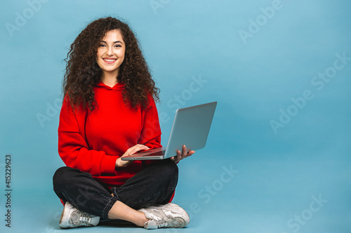 Quadro su tela Happy young curly beautiful woman sitting on the floor with crossed legs and using laptop isolated on blue background