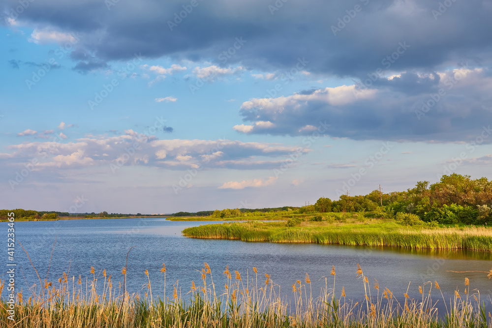 Beautiful river coast at sunset in summer. Colorful landscape with lake, green trees and grass, blue sky with multicolored clouds and orange sunlight reflected in water.