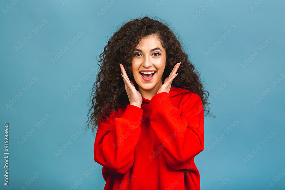 Happy cheerful young woman with curly hair rejoicing at positive news or birthday gift, with joyful and charming smile. Student girl relaxing after college isolated over blue background.