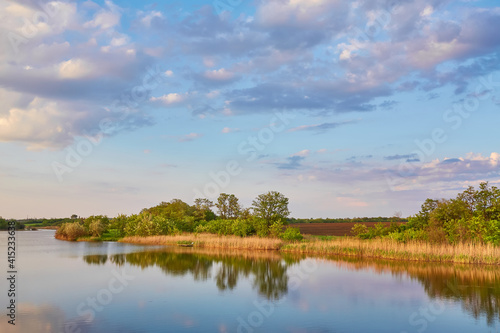 Beautiful river coast at sunset in summer. Colorful landscape with lake, green trees and grass, blue sky with multicolored clouds and orange sunlight reflected in water.